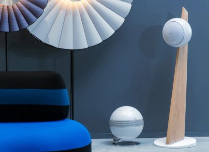 Hvid designerhøjttaler på fod i lyst træ med en anden hvid sfærisk model i en moderne blå og sort stue