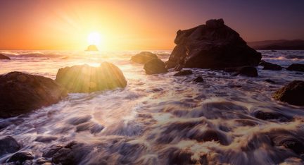 Rochas em um mar agitado ao pôr do sol, ondas espumantes, céu laranja e amarelo brilhante, sol baixo no horizonte
