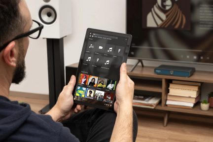 Barzdotas vyras, laikantis planšetę su muzikos programėle, balta kolonėlė ant stovo, televizorius, baldai su knygomis ir prietaisas