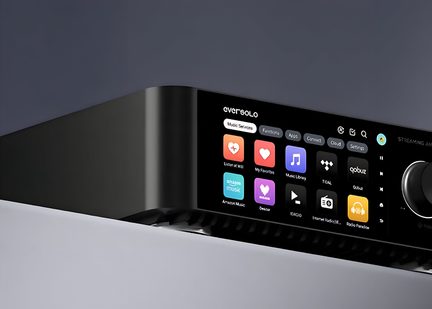 Close-up three-quarter front view of a rectangular black metal electronic device, featuring a color touchscreen displaying music service icons and a large side rotary knob