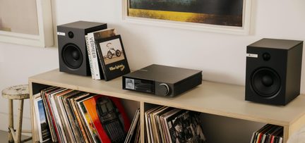 Impianto hi-fi con amplificatore nero metallizzato dotato di display e di una grande manopola frontale, affiancato da due diffusori da scaffale neri opachi con altoparlante circolare, visto da tre quarti leggermente dall’alto, il tutto posizionato su una mensola in legno chiaro piena di dischi in vinile