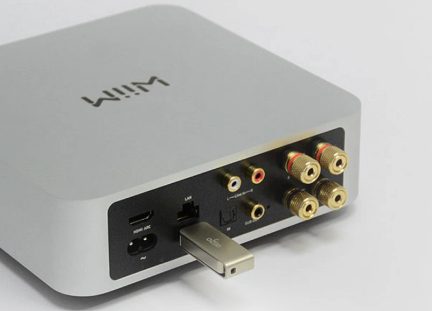 Pravokotni siv ohišje s priključki za avdio, HDMI, LAN, USB in RCA na zadnji strani, USB ključek vstavljen v USB priključek