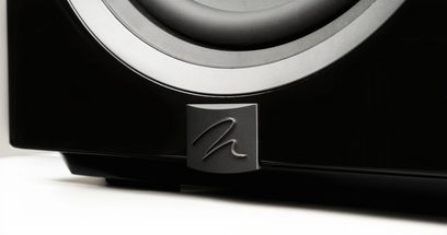 Dynamo 12 subwoofer i sort lak, delvis set fra en lav vinkel, skinnende sort overflade, metallisk logo i relief, synlig cirkulær højttalerkant