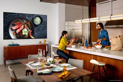 Samsung QE65Q900R-tv, der viser en tallerken med grillet kød i et moderne køkken med to personer, der laver mad