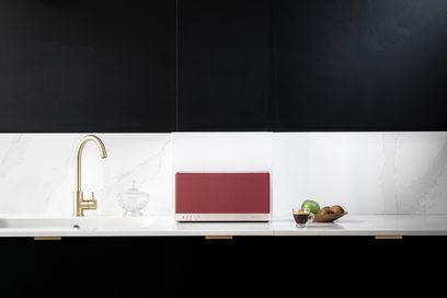 Connected wireless Triangle AIO 3 speaker in red ochre on white marble countertop with golden faucet, fruit bowl, and mug