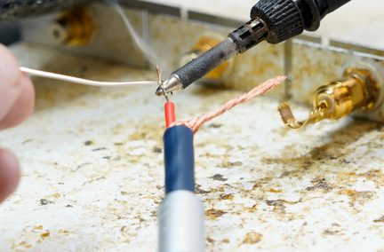 Primo piano in vista obliqua di un saldatore nero con punta metallica argentata applicato su un conduttore rosso e una treccia di rame di un cavo coassiale con guaina blu su un piano di lavoro macchiato, leggero fumo che sale