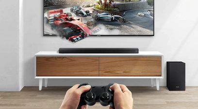 Samsung HW-R530 sort soundbar under et TV, der viser en scene fra et bilspil, med en spillekonsol i forgrunden