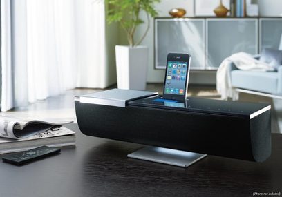 Sort Onkyo ABX-100 dockingstation med iPhone placeret på den, set forfra på et træbord med magasiner og en plante i baggrunden