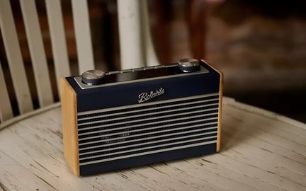 Retro rektangulär bärbar radio, sedd från tre fjärdedels framifrån, front i mörkblå metallisk horisontell gallerdesign, ljus träpanel på sidorna, två stora silverfärgade rifflade knappar på ovansidan