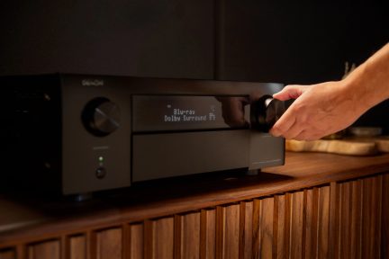 Hlavním prvkem je audio-videový přijímač Denon Blu-ray s Dolby Surround, umístěný na dřevěné polici.