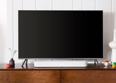 Hvid Sonos Beam lydbjælke, set forfra, placeret på et træ-TV-møbel, under en stor slukket TV-skærm