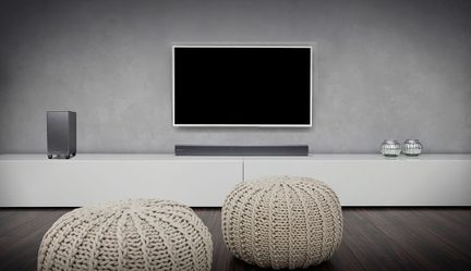 Fladskærm, soundbar, to runde strikkepoffer i beige på mørkt trægulv foran lys gråmur