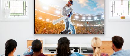 Mand, der spiller cricket på en overfyldt stadion, set bagfra på en tv-skærm foran et siddende publikum