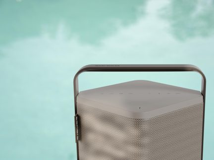 Altavoz Bluetooth portátil rectangular beige con gran asa metálica, vista de cerca al borde de una piscina