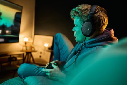 Een man in profiel met een zwarte koptelefoon, die een gamecontroller vasthoudt, zittend voor een scherm en een brandende lamp.