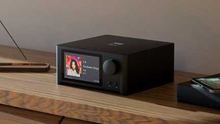 Rectangular black case with color screen, circular button, 2 buttons at the bottom right, placed on a wooden surface