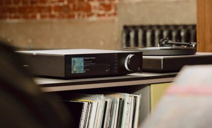 Amplificatore audio nero con schermo a colori e grande manopola rotante, posizionato su un mobile accanto a un giradischi