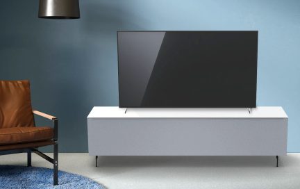 Vypnutá černá televize na bílém nábytku před modrou zdí, hnědé křeslo vlevo, šedá závěsná lampa nad ní