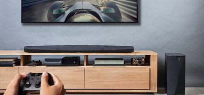 Czarna listwa dźwiękowa Denon DHT-S716H pod ekranem wyświetlającym samochód wyścigowy, z pilotem do gier i urządzeniami audio
