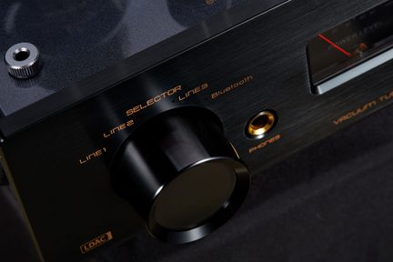 Nærbillede i trekvart vinkel af et sort børstet aluminiums frontpanel på en lydafspiller, med en stor cylindrisk sort vælgerknap omgivet af orange markeringer, hovedtelefonudgang med guldkant og delvist synligt VU-meter