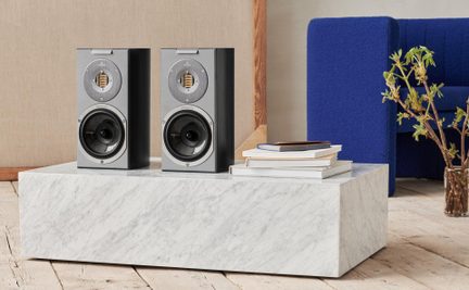 To rektangulære højttalere i mat sort med front i sølvmetal, hver udstyret med en gylden bånddiskant og en sort glas-woofer, set forfra, placeret side om side på en lav blok af hvid marmor med årer