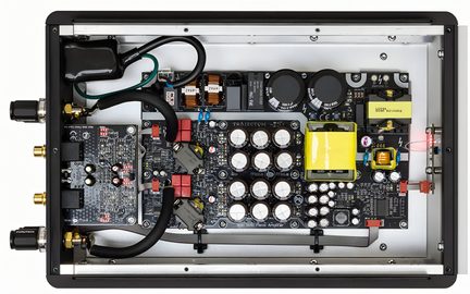 Vista superior de un amplificador de audio abierto, caja rectangular de metal negro y gris que contiene placas de circuito impreso negras densamente pobladas, condensadores cilíndricos blancos, conectores RCA dorados laterales y transformador amarillo aislado