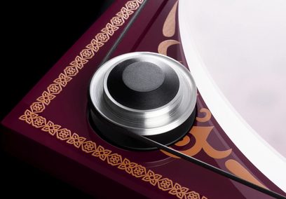 Hi-fi pladespiller Pro-Ject Essential III George Harrison, nærbillede af en cirkulær sølv- og sort pladevægt med dekorativ gylden kant på bordeaux baggrund, tynd sort kabel synlig