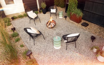 Gravel patio seen from above, four black cord chairs with white cushions around a lit metal fire pit