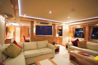 Luksuriøst yachtinteriør med beige sofaer, røde puder, trærammer, indbyggede TV-skærme og integreret loftbelysning