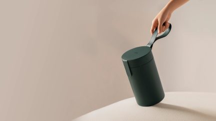 Hoved, der holder en cylindrisk grøn skraldespand med låg og håndtag, set fra siden, ensfarvet beige baggrund