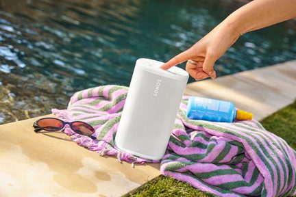 Hvid Sonos bærbar højttaler ved poolkanten, placeret på et stribet håndklæde med solbriller og solcreme