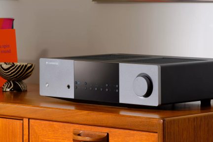 Črno-siv stereo ojačevalnik Cambridge AXA na lesenem regalu, sprednja plošča z gumbi in velikim gumbom za glasnost