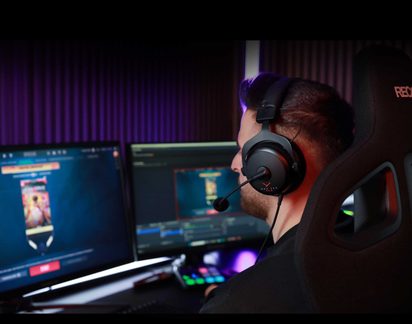 Mann i profil med svart hodetelefon, sittende på en svart gamingstol, foran 2 dataskjermer, lilla bakgrunn