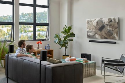 Living modern, vedere oblică, canapea din piele gri, televizor montat pe perete, bară de sunet neagră, boxe pe stative, măsuță de cafea din sticlă