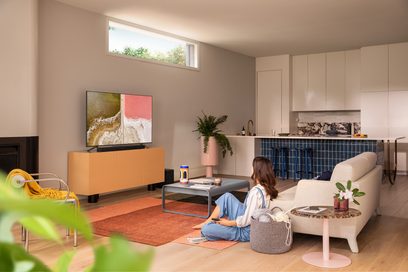 Moderne woonkamer met beige bank, salontafel, tv-meubel, oranje tapijt, groene plant en een televisie waarop een strand en golven te zien zijn