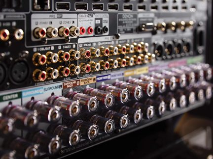 Detailní šikmý záběr na zadní kovovou černou stranu audio zesilovače, vybavenou řadami kovových RCA konektorů pozlacených s červenými a bílými kroužky a svorkami pro reproduktory z průhledného plastu