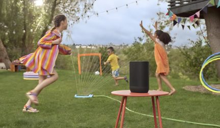 Altavoz negro sobre mesa roja, niños jugando con rociador en césped verde, guirnalda de banderines coloridos
