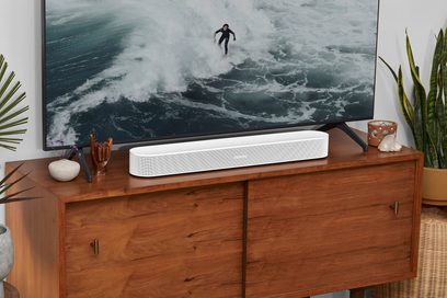 Biała matowa listwa dźwiękowa Sonos Beam (Gen 2) na drewnianej szafce, pod telewizorem wyświetlającym surfera na potężnej fali