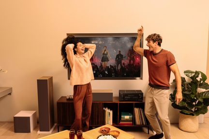 Hjemmebiografsæt med gulvhøjttalere og subwoofer foran vægmonteret tv i en stue med et dansende par