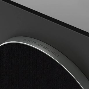 Částečný úhlový pohled na subwoofer Sira L-1, zakřivený stříbrný okraj s nápisem, černý texturovaný povrch, šedé pozadí