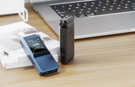 Kaks ristkülikukujulist metallist kaasaskantavat heliseadet: üks sinine, anodeeritud ja lamavas asendis, ekraanil kiri „LDAC” ning topelt USB-port; teine tumehall, tekstuurse pinnaga, püstises asendis, kolmekvartali vaates, helitugevuse rulliku ja nupureaga. Mõlemad asetsevad puidust laual sülearvuti kõrval.