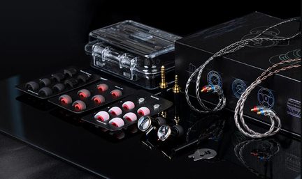 Slightly top-down view of a set of silver metal in-ear headphones with a silver-copper braided cable, accompanied by numerous black and red silicone ear tips displayed on black plates, two gold-plated jack adapters, and a transparent carrying case, all placed on a glossy black surface