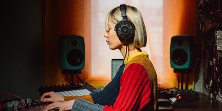 Sort circum-aural hovedtelefon i plastik og perforeret metal, set fra siden, båret af en person i et musikproduktionsmiljø