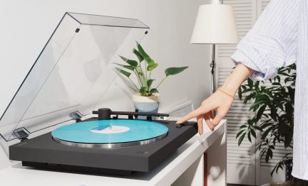 Sort pladespiller med gennemsigtigt låg, turkis vinylplade, hånd der justerer afspilningen på hvidt møbel i moderne stue