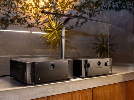 Dos equipos de audio hi-fi negros de metal, con frontales cepillados, pantalla redonda central y botones grandes, vista en tres cuartos.