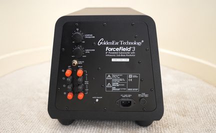 Vista traseira de um subwoofer preto com controles, conectores vermelhos e pretos, inscrições técnicas e entrada de alimentação.