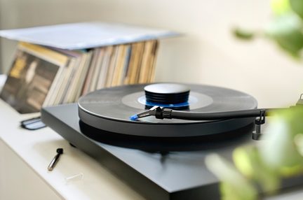 Černá vinylová deska s otáčejícím se diskem, rameno s modrou jehlou, v pozadí police s deskami