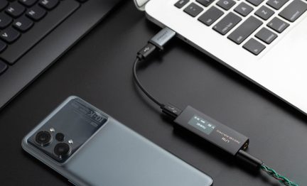 Topvinkel, lille sort audio-konverter i aluminium med OLED-skærm, USB-C-port og jack-udgang forbundet med kabel til en sølvfarvet bærbar computer, ved siden af en satinblå smartphone med triple kamera på sort skrivebord
