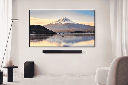 Avskalad vardagsrumsmiljö sedd framifrån med en väggmonterad TV med tunn svart ram som visar ett landskap, ovanför en rektangulär svart soundbar med matt finish och en kubformad svart subwoofer placerad på golvet, ljusa väggar, fåtölj i beige tyg och minimalistiska dekorativa inslag