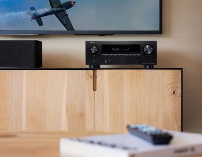Ampli home-cinéma Denon AVR-X2800H DAB Noir, vue de face, télécommande sur meuble bois clair, avion militaire sur écran TV